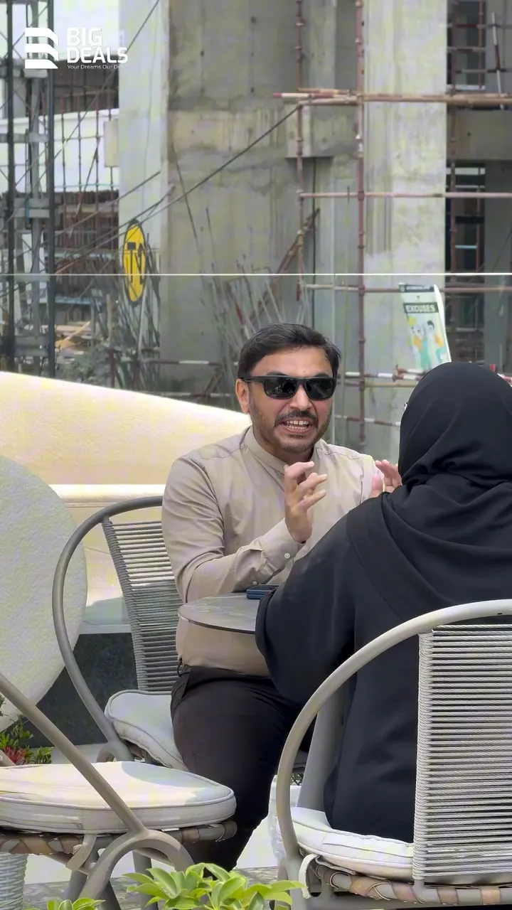 A presenter and a visitor over coffee on a terrace, with a building under construction behind them, in Big Deals' Prestige Ocean Pearl film
