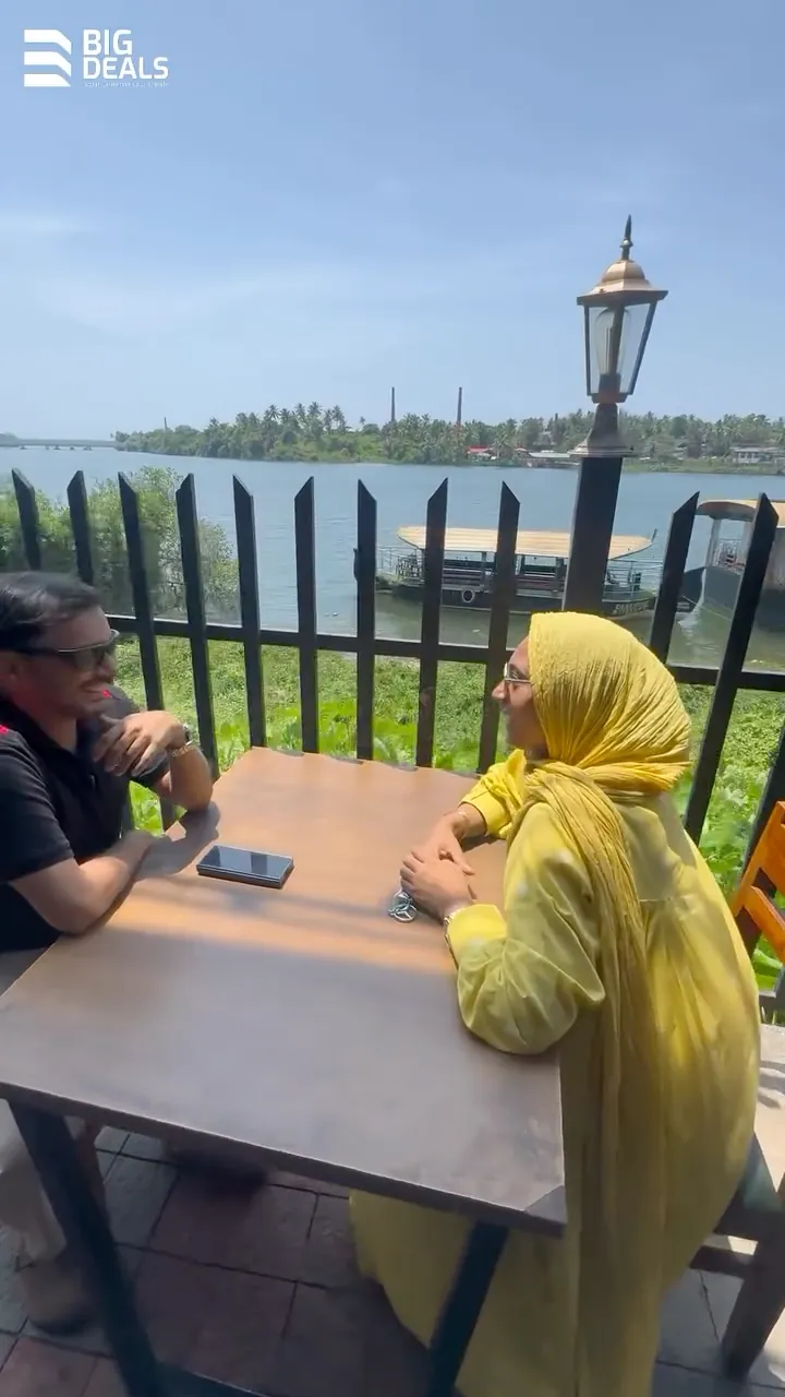 Two presenters at a café table beside the river in Feroke, at the start of Big Deals' Sobha Bellevue film