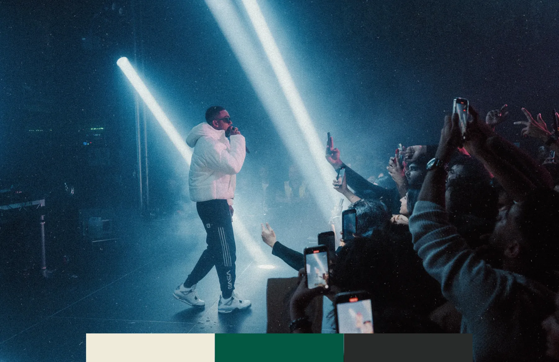 The artist in a white puffer under stage-light beams, phones raised in the crowd, with the three brand colours as a band along the foot.