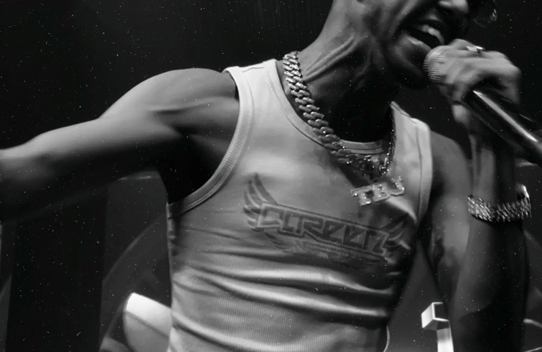 A black-and-white close-up on stage: mic in hand, a TBJ pendant on the chain.