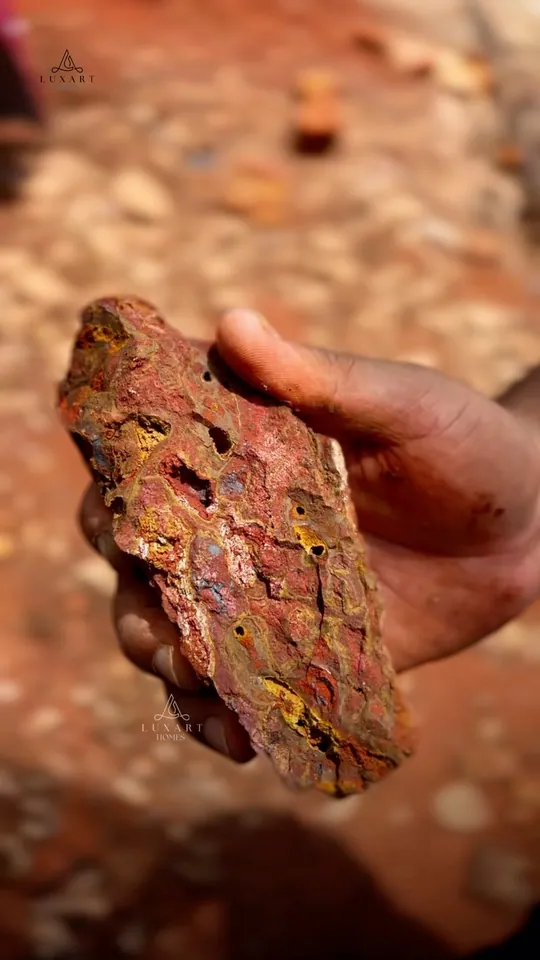 A hand holding a rough red laterite stone, on the cover of a Luxart Builders explainer reel
