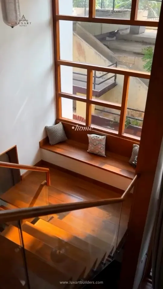 A wooden window seat under a tall window beside a glass-railed staircase, from a walkthrough reel on the Luxart Builders website